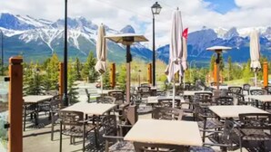 An outdoor café with tables and chairs, umbrellas, and a mountain backdrop. - Mountain Getaway in Canmore! Sleeps 6 - Pool & Hottub (Canmore)