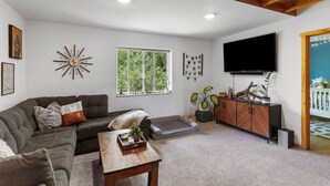 A living room with a sectional sofa, a coffee table, a TV, and a window with a view of greenery. - Beautiful Home on River - 30 Day Rental Discounts! (Steamboat Springs)