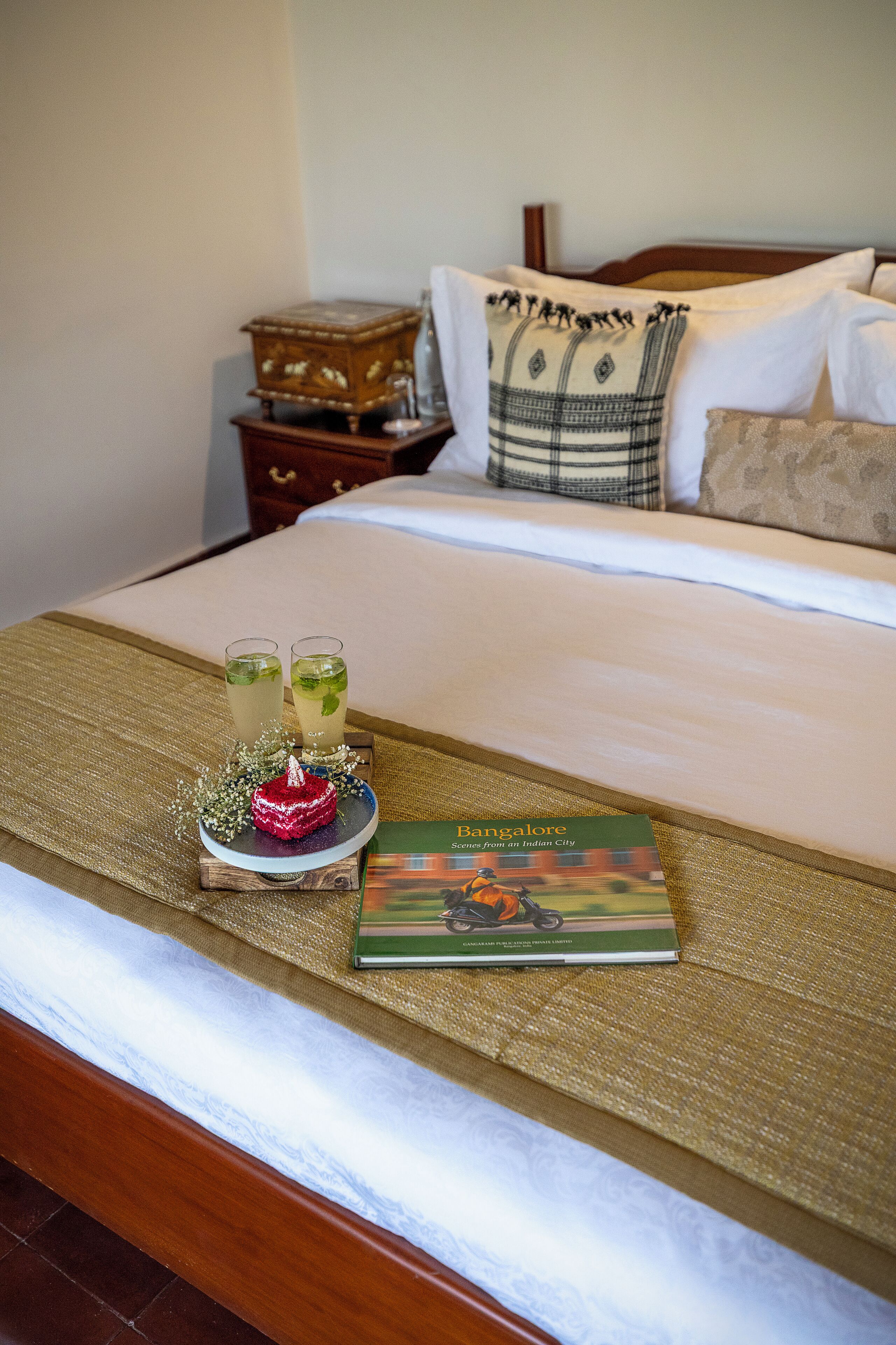 A neatly made bed with a book titled 'Bangalore' and two glasses of a beverage on the bedspread.