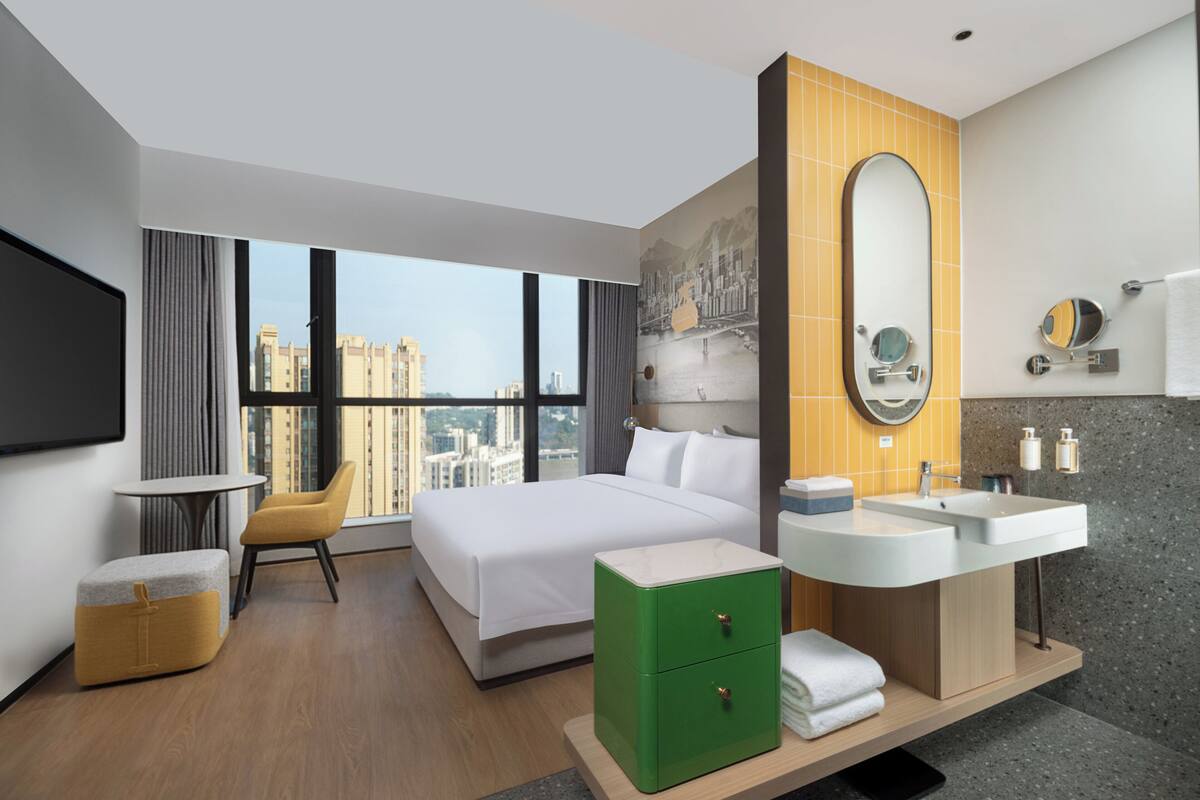 A modern hotel room with a large bed, a green nightstand, a bathroom with a large mirror, and a city view through the window.