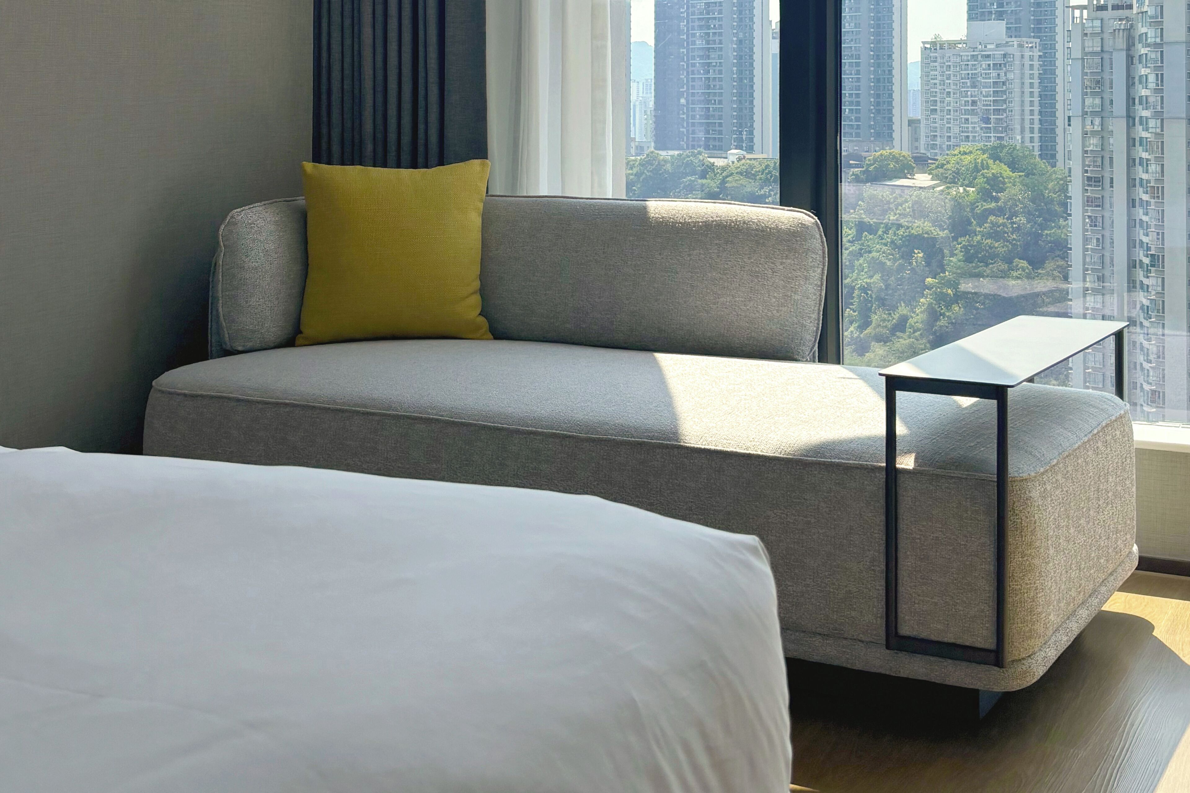 A modern hotel room with a grey sofa, a small side table, and a large bed.