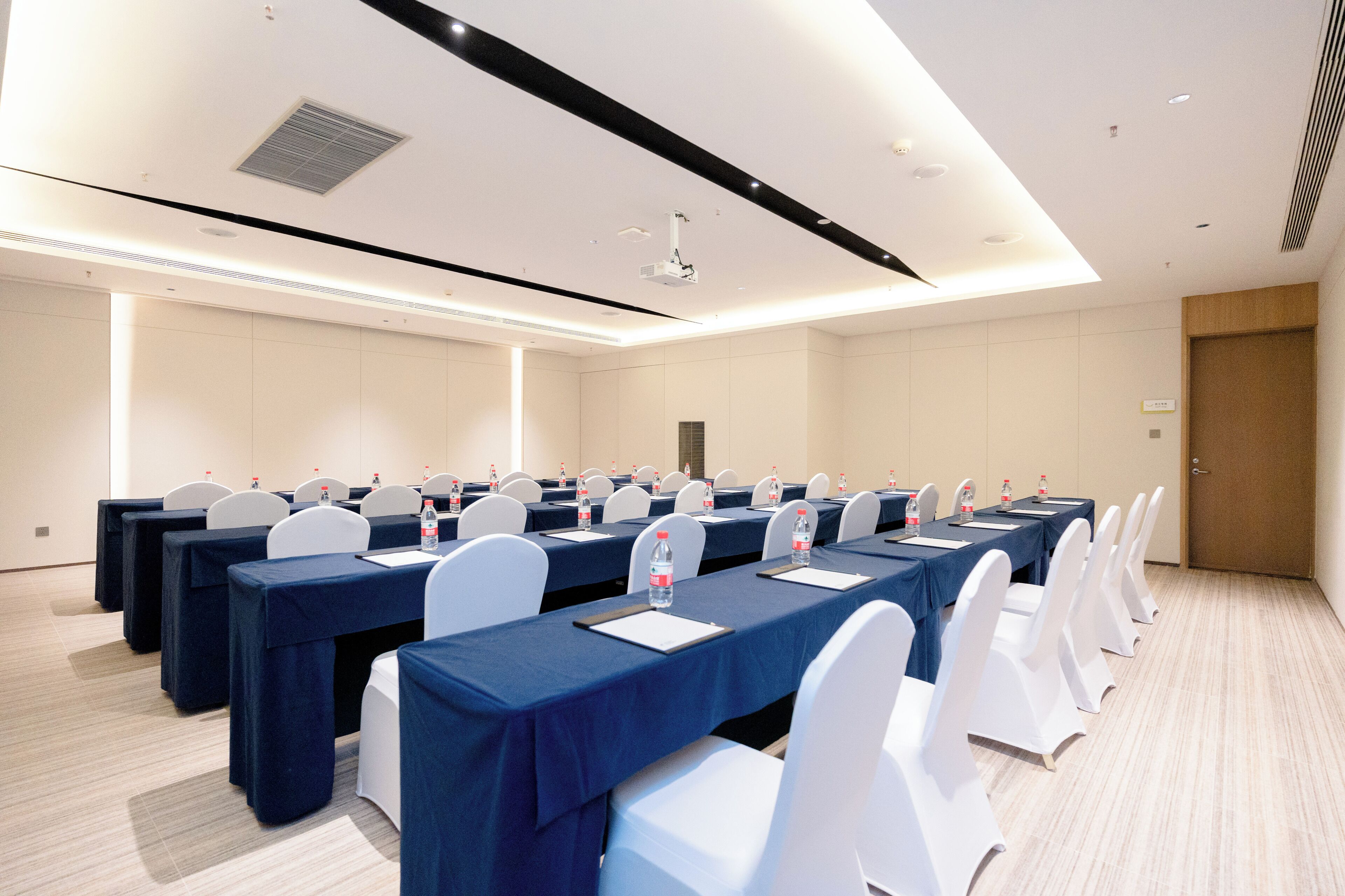 A conference room with rows of tables, white chairs, and a projector on the ceiling.