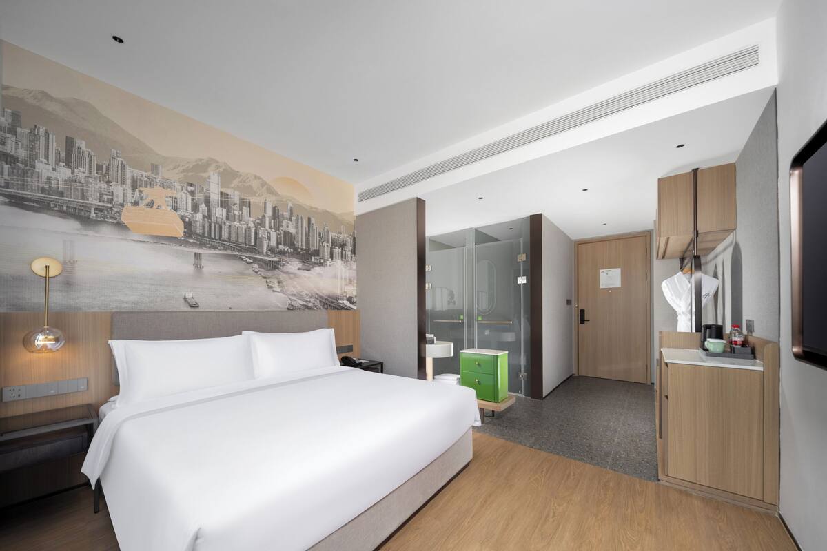 A modern hotel room with a large bed, a cityscape mural, a walk-in closet, and a small kitchenette.