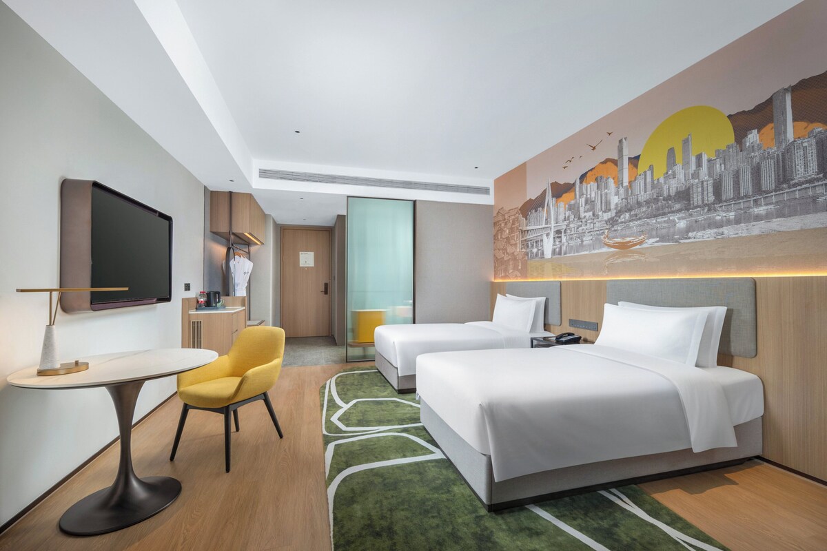 A hotel room with two beds, a desk, a chair, a TV, and a painting of a cityscape.
