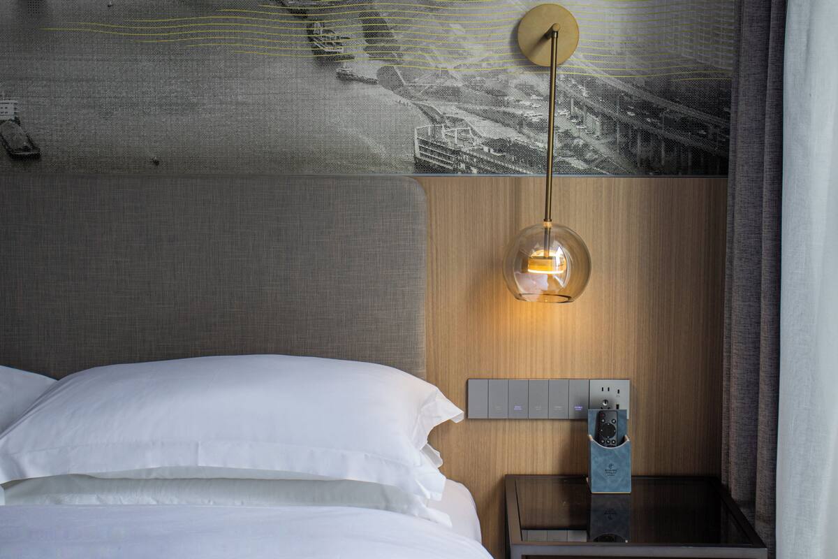 A hotel room with a bed, a nightstand with a lamp, and a wall-mounted control panel.