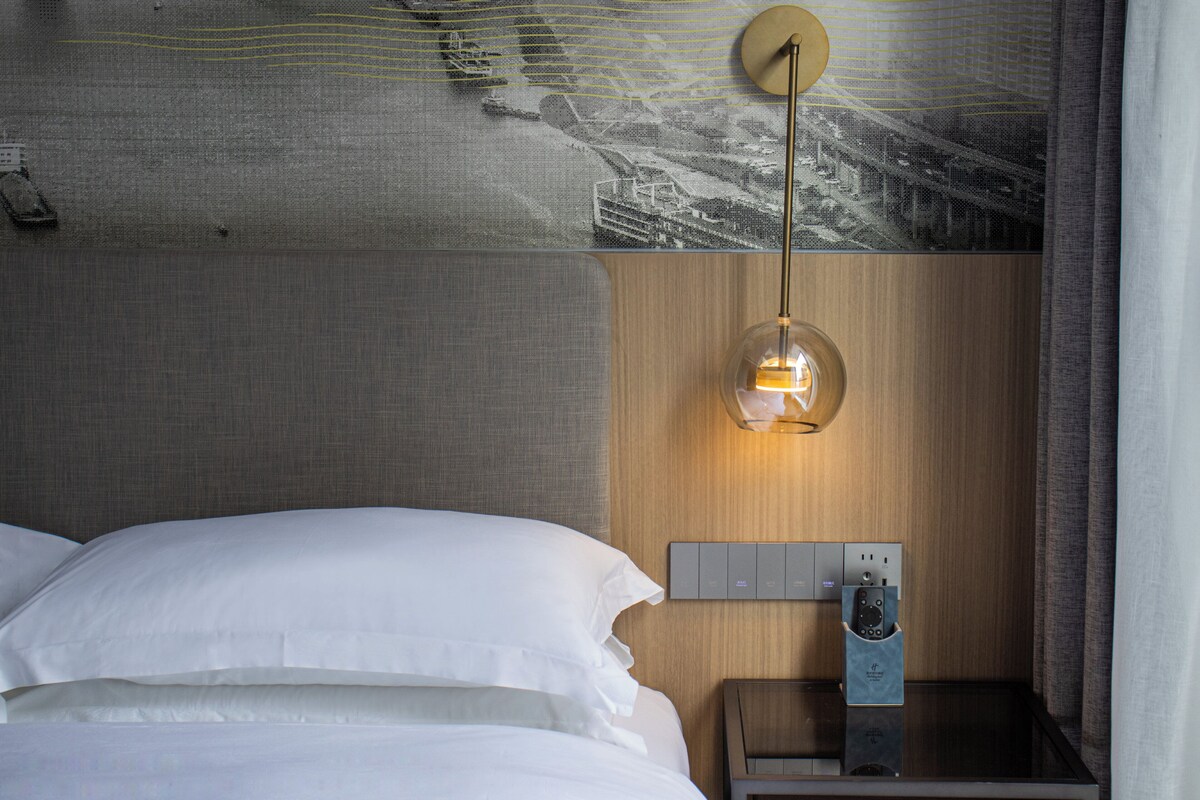 A hotel room with a bed, a nightstand with a lamp, and a wall-mounted control panel.