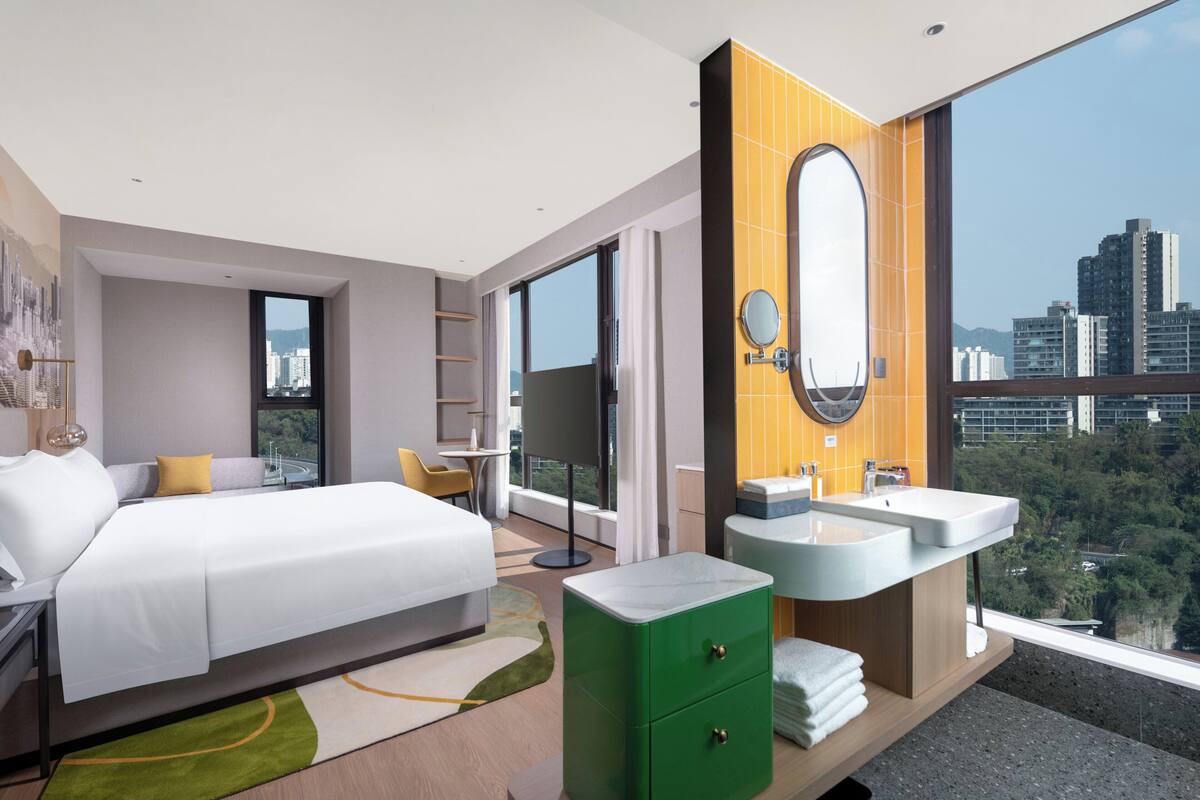 A modern hotel room with a large bed, two sinks, a mirror, and a view of the city.