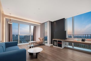 Una sala de estar moderna con un televisor grande, un sofá, una mesa de centro y vistas de la ciudad a través de grandes ventanales.