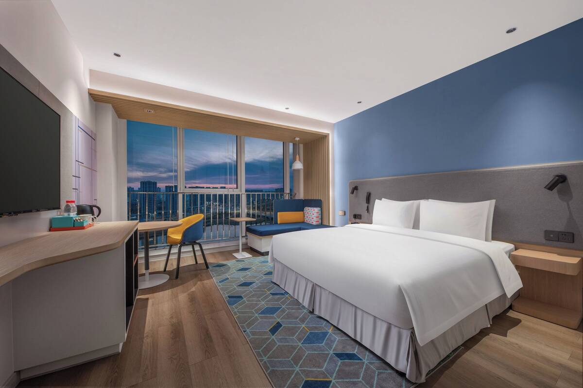 A modern hotel room with a large bed, a desk, a chair, and a view of the city through large windows.