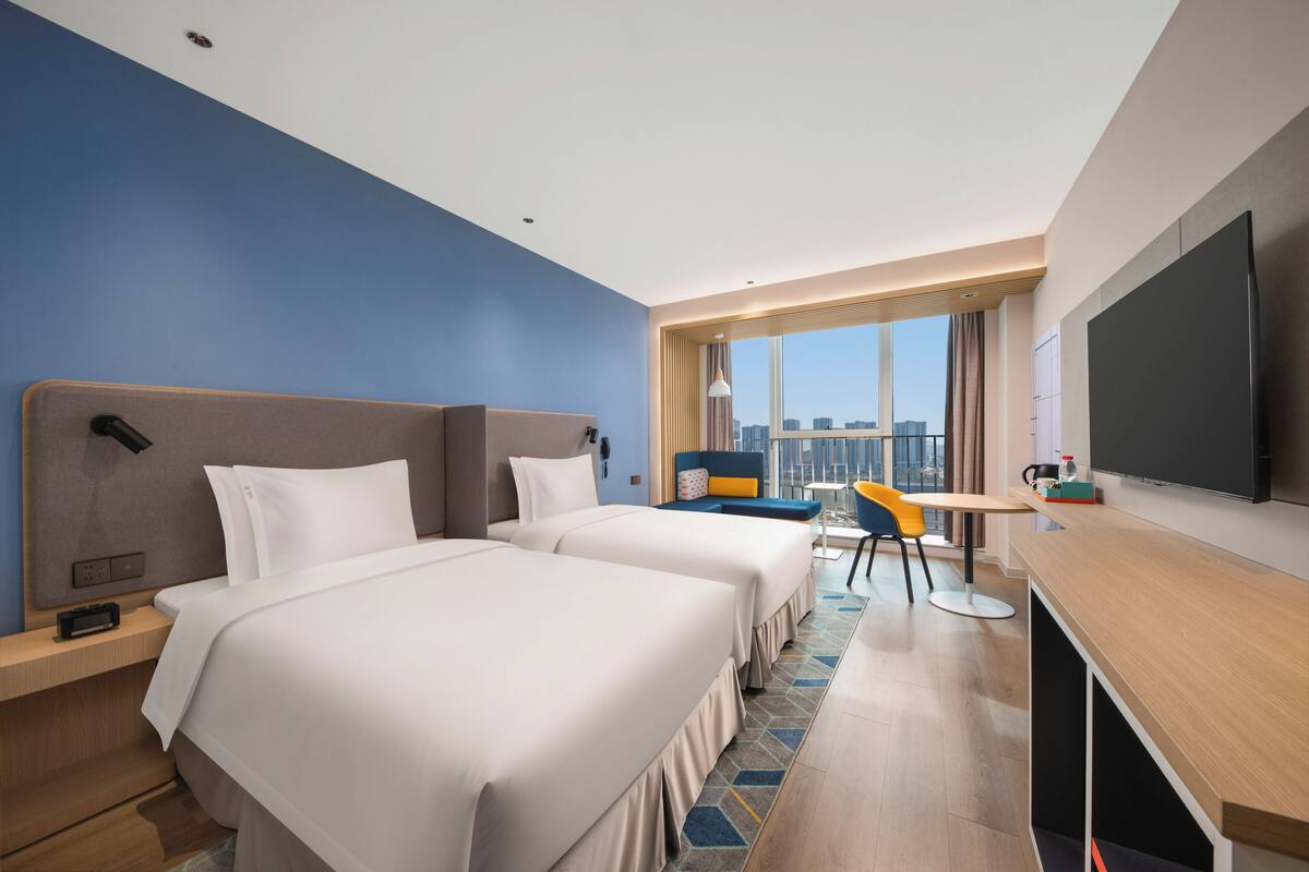 A hotel room with two beds, a desk, a TV, and a city view.