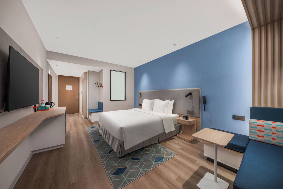 A modern hotel room with a large bed, a desk, a flat-screen TV, and a seating area with a blue cushion.