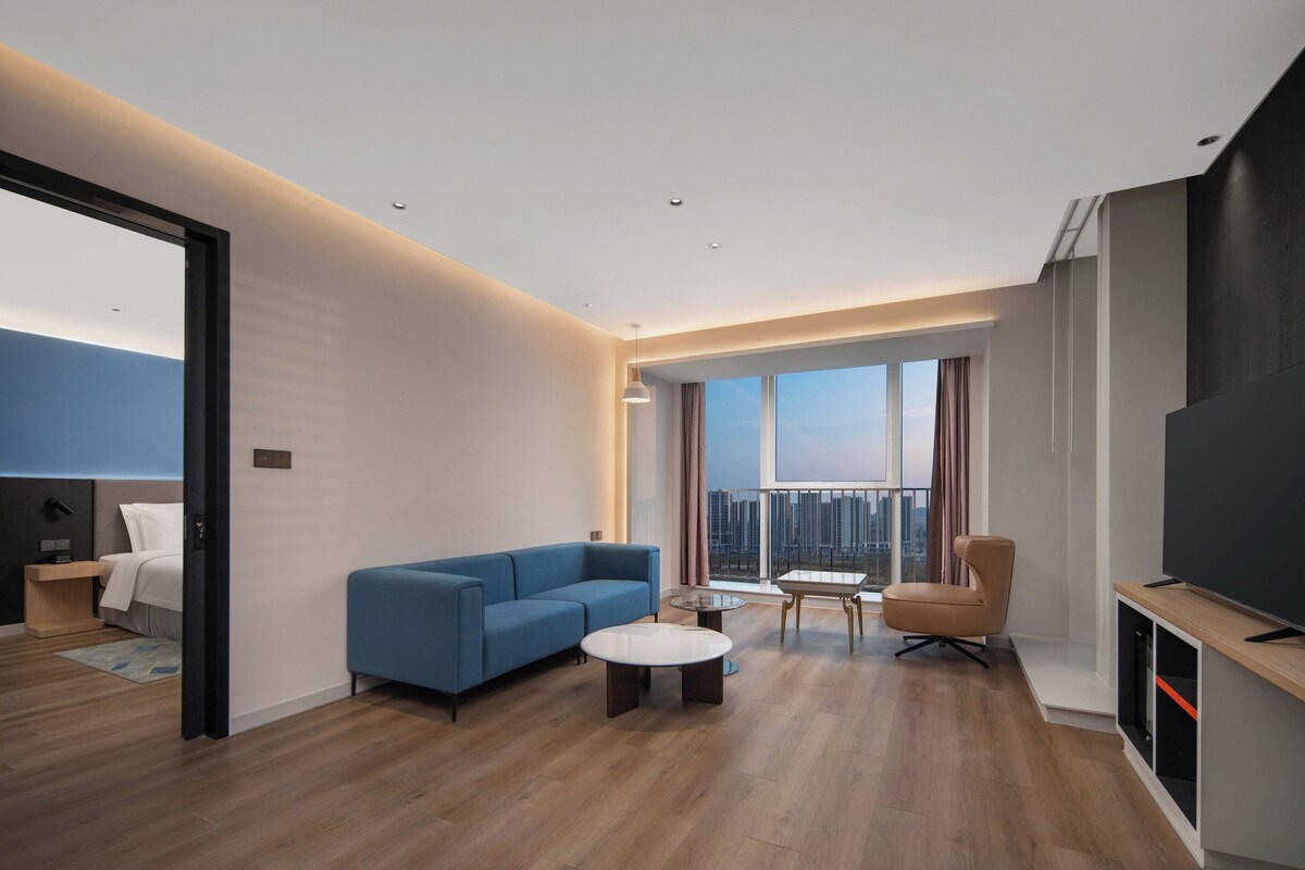 Una camera d'hotel moderna con un divano blu, un piccolo tavolino rotondo, una TV e un'ampia finestra con vista sulla città.