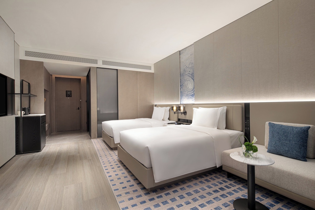 A modern hotel room with two beds, a small table with a lamp, a sofa, and a TV.