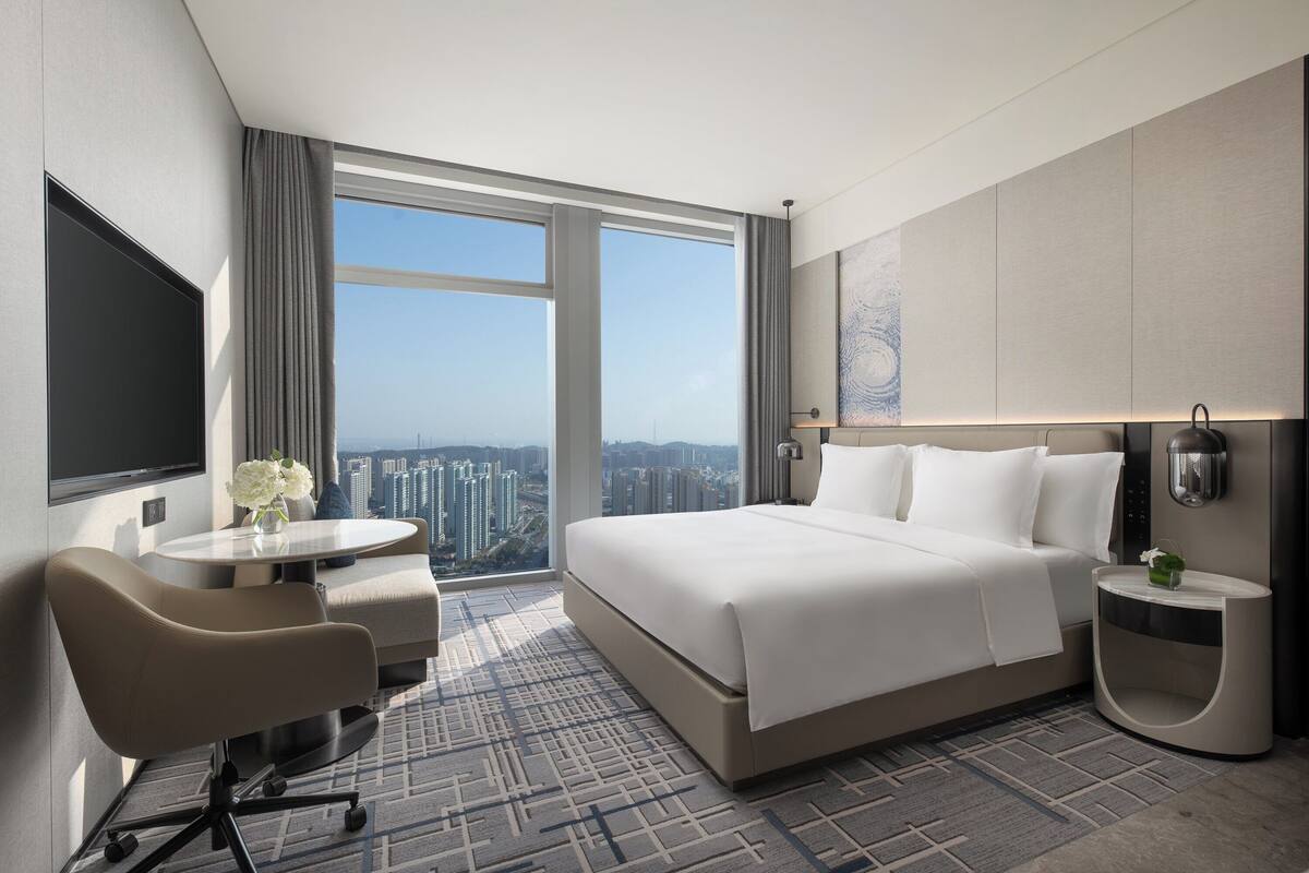 A modern hotel room with a large bed, a desk, and a view of the cityscape.