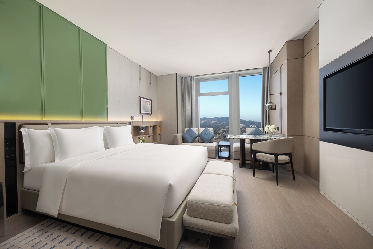 Ein modernes Hotelzimmer mit einem großen Bett, einem Schreibtisch, einem Sessel und einem Fernseher.