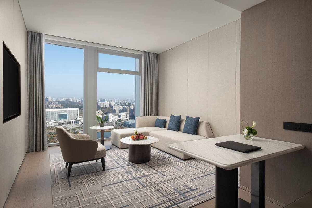 A modern hotel room with a large window offering a city view, a sofa, a chair, a small table, and a flat-screen TV.