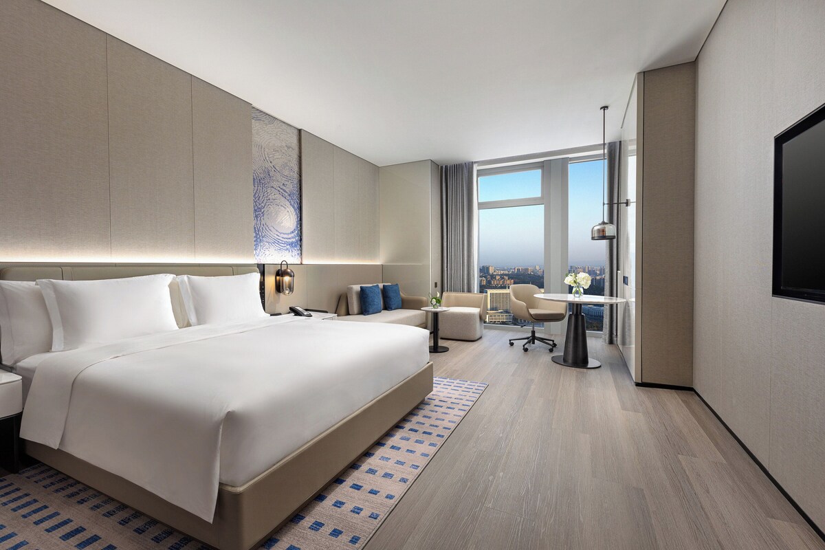 Ein modernes Hotelzimmer mit einem großen Bett, einem Schreibtisch, einem Sessel und Blick auf die Stadt.