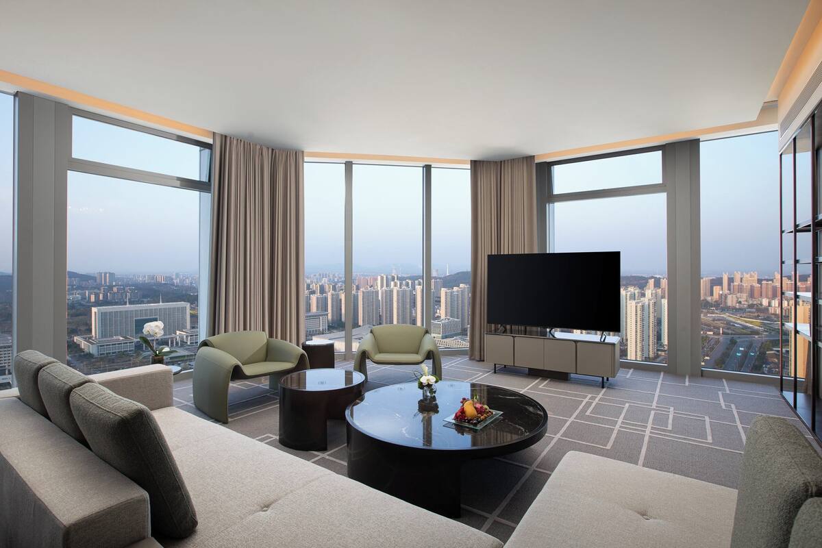 A modern hotel room with a large city view, a flat-screen TV, and a comfortable seating area with a coffee table.
