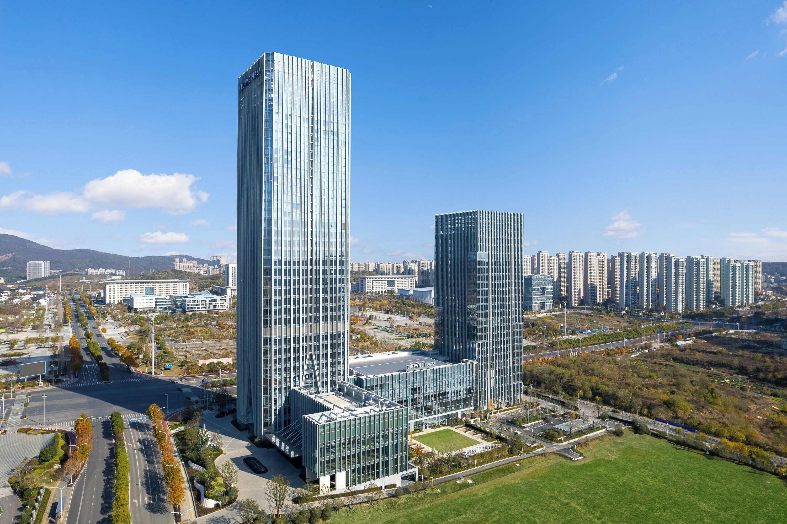 A modern high-rise building complex with a glass facade, surrounded by a well-maintained park and a road.