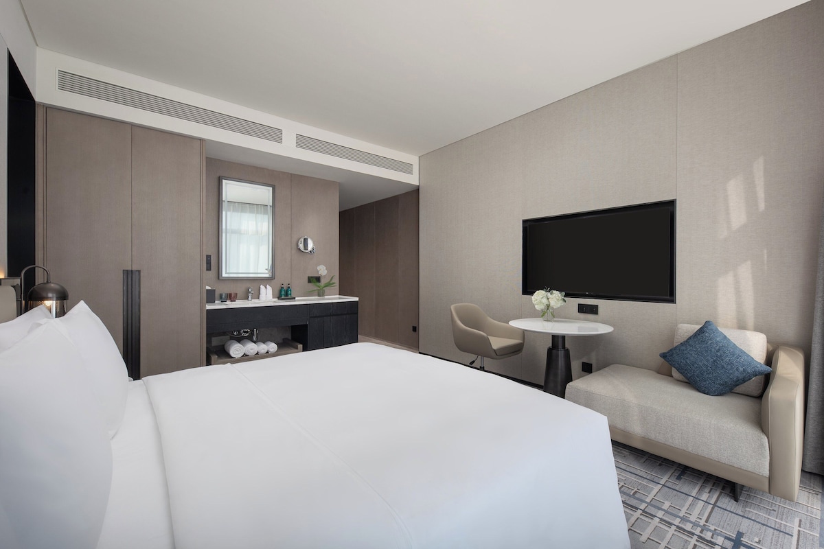 A modern hotel room with a large bed, a desk with a chair, a small sofa with a pillow, a TV mounted on the wall, and a window with curtains.