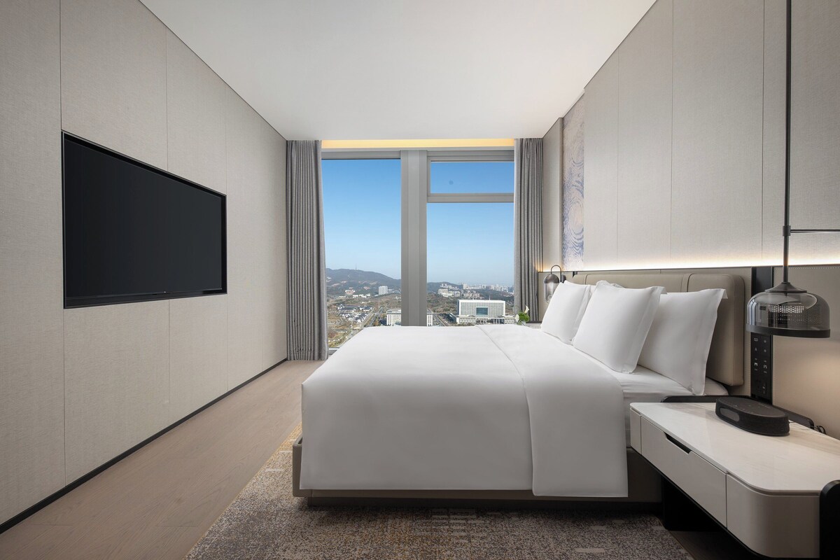 A modern hotel room with a large bed, a flat-screen TV mounted on the wall, a bedside table with a lamp, and a view of the city through large windows.