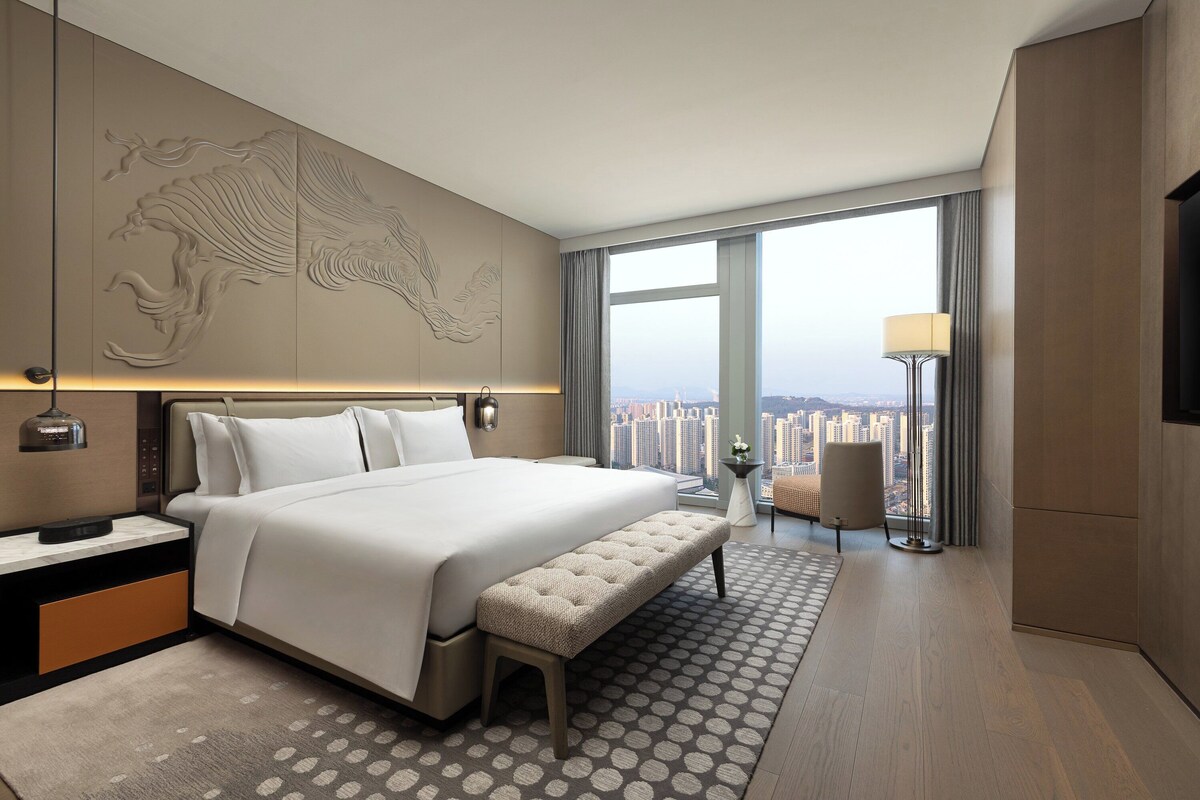 Ein modernes Hotelzimmer mit einem großen Bett, einer Sitzbank, Blick auf die Stadt und einer dekorativen Wandverkleidung.