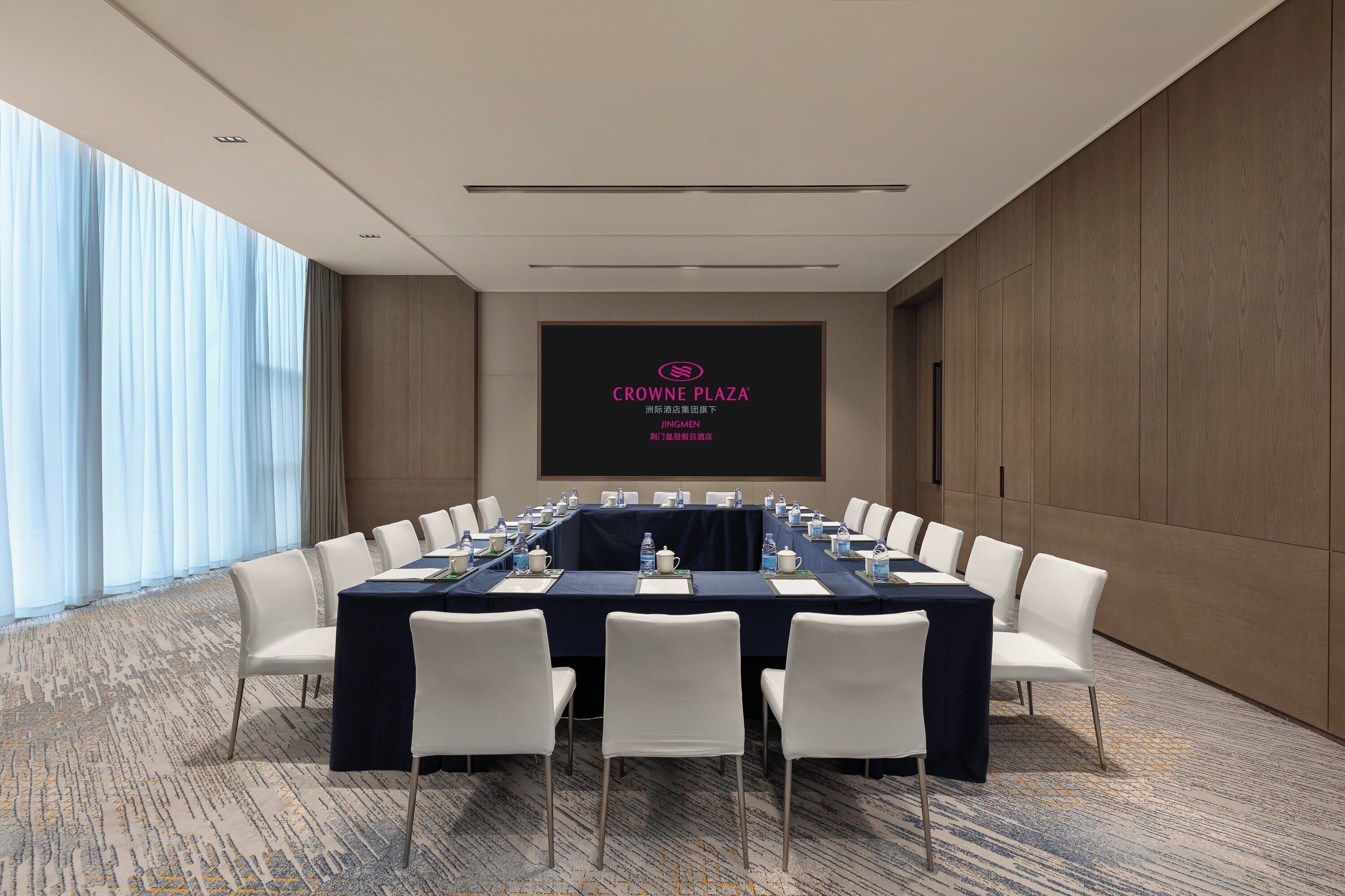 A conference room with a long table, white chairs, and a screen displaying "CROWNE PLAZA JINGMEN".