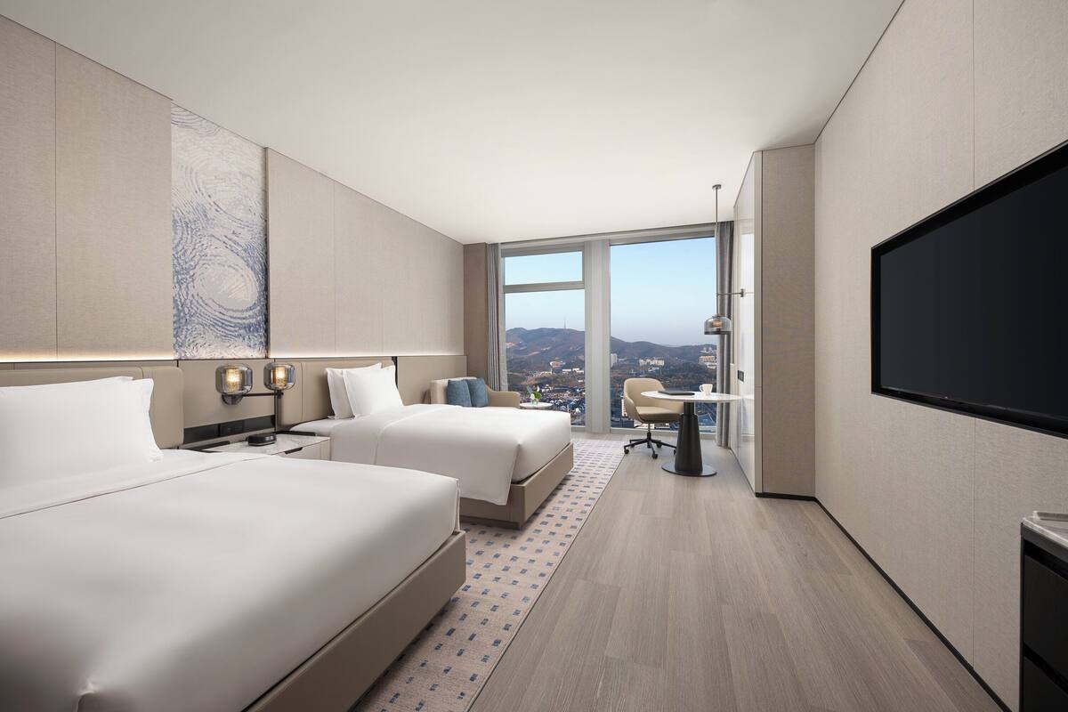 A modern hotel room with two beds, a desk, a chair, and a large TV screen.