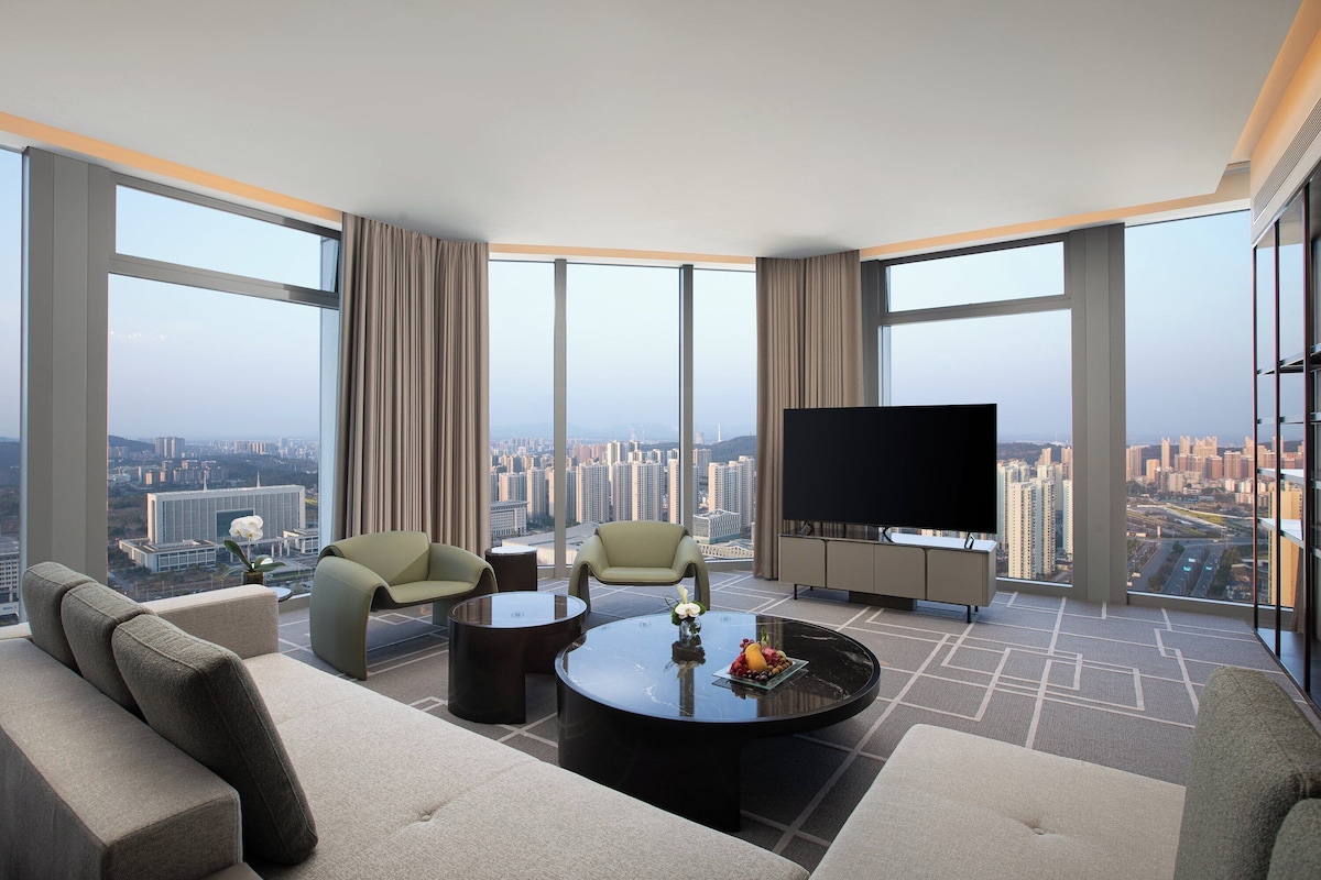 A modern hotel room with a large city view, a flat-screen TV, and a comfortable seating area with a coffee table.