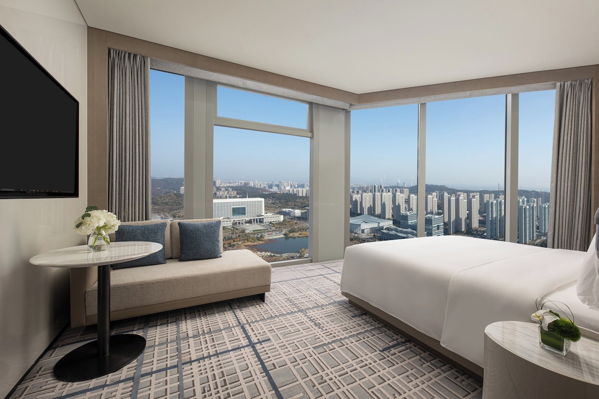 A hotel room with a large window, a bed, a sofa, a small table, and a TV.