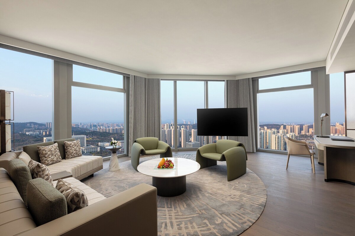 A modern living room with a sofa, two armchairs, a coffee table, and a flat-screen TV. The room features large windows offering a city view.