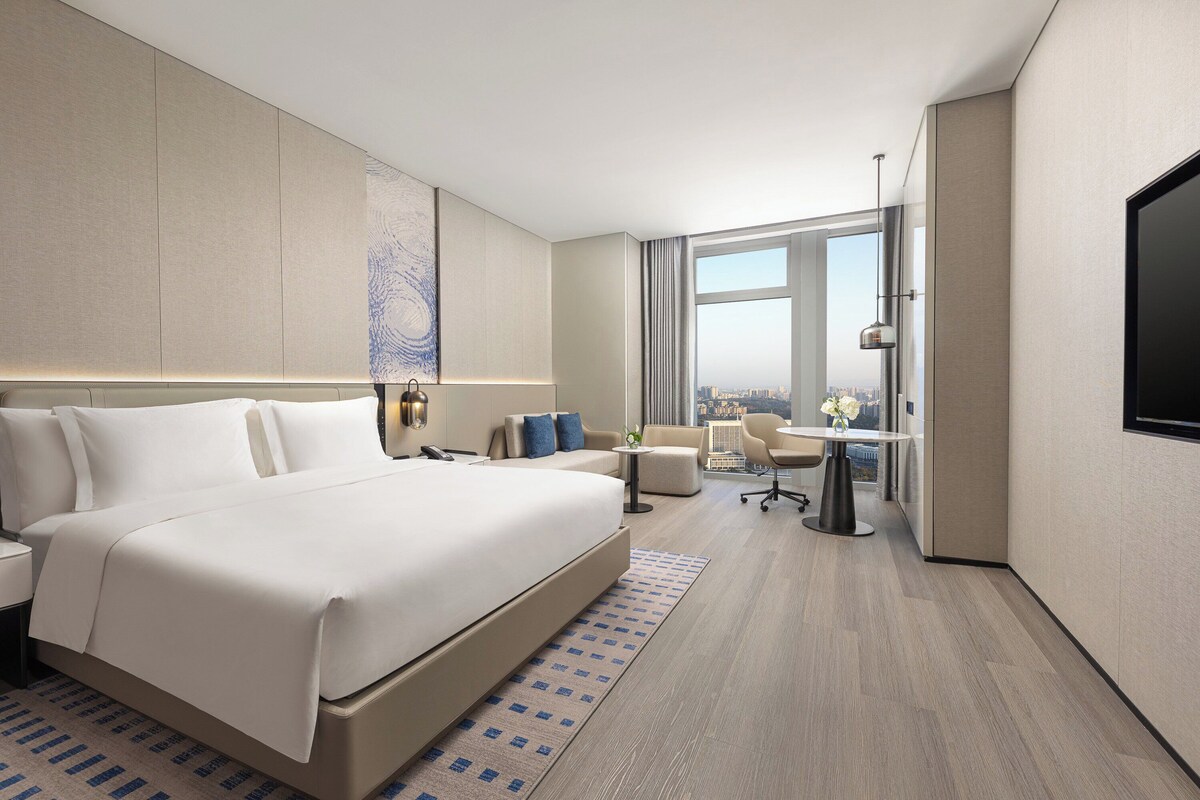 Ein modernes Hotelzimmer mit einem großen Bett, einem Schreibtisch, einem Sessel und Blick auf die Stadt.