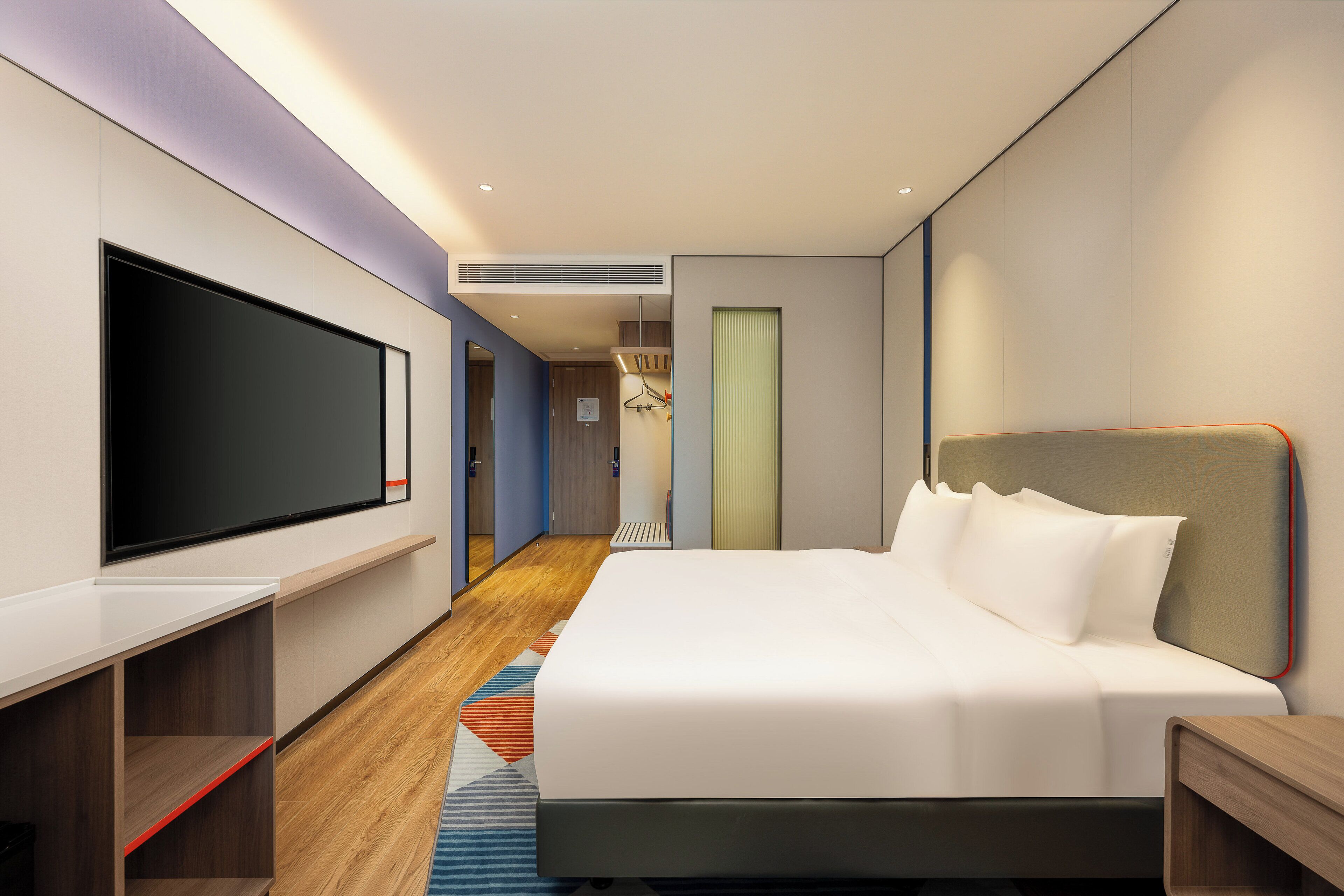 A modern hotel room with a large flat-screen TV, a bed with white linens, a wooden floor, and a built-in shelf.