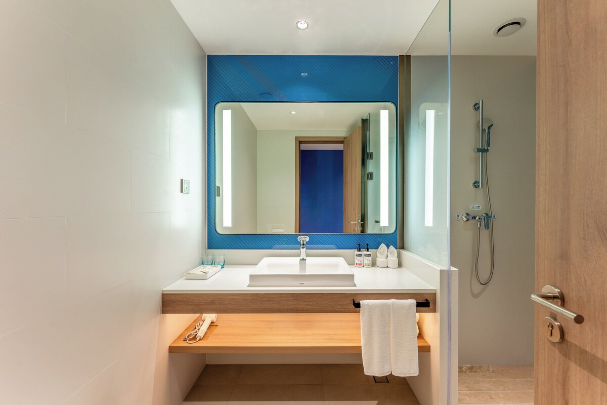 A modern bathroom with a large mirror, a sink, a shower, and a wooden bench.