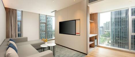 Una sala moderna con un sofá, un televisor de pantalla plana, una pequeña mesa con un florero y ventanas amplias con vista a la ciudad.