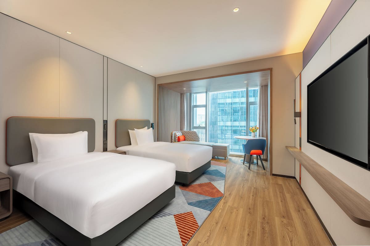 Habitación de hotel con dos camas, un televisor grande y vista a la ciudad.