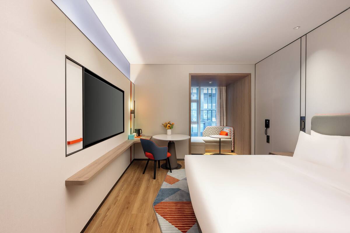 Una habitación de hotel moderna con una cama grande, un escritorio con silla, una mesita redonda con un jarrón de flores y vistas a la ciudad a través de amplios ventanales.