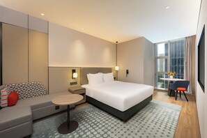 Une chambre d’hôtel moderne, dotée d’un grand lit, d’un canapé, d’une petite table et offrant une vue sur la ville grâce à une grande fenêtre.