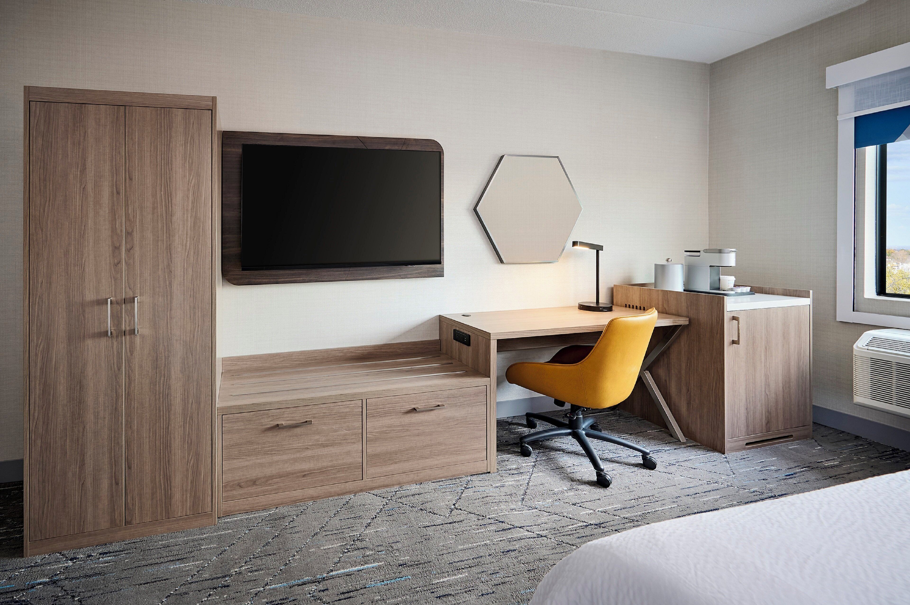A hotel room with a wooden desk, a chair, a bed, a wardrobe, a flat-screen TV, and a window with a view.