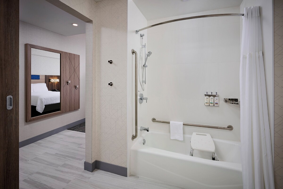 A modern bathroom with a bathtub, shower, and a mirror with a bed in the background.
