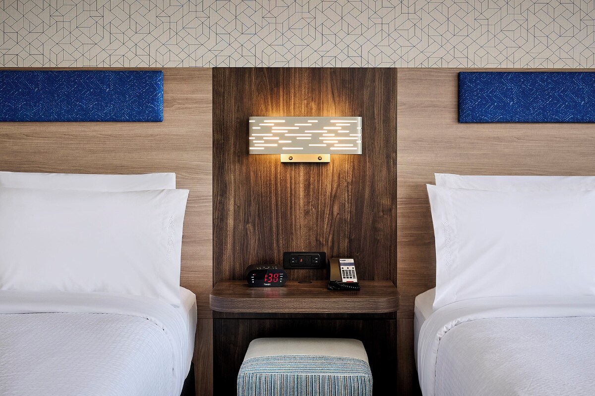 A hotel room with two beds, a bedside table with a digital clock, a telephone, and a wall-mounted light fixture.