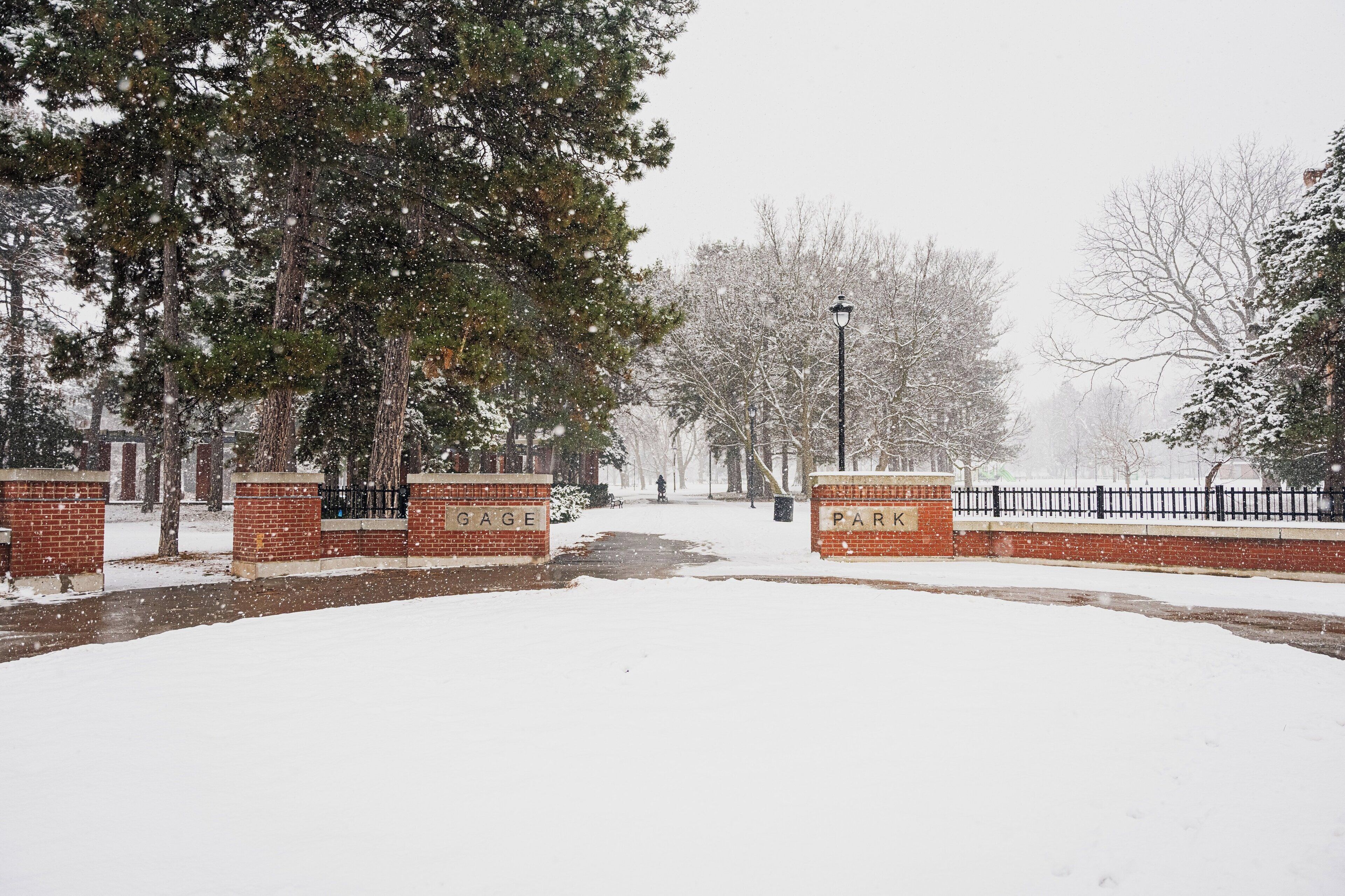 A snow-covered park with a brick wall labeled 'GAGE PARK' and a metal fence.