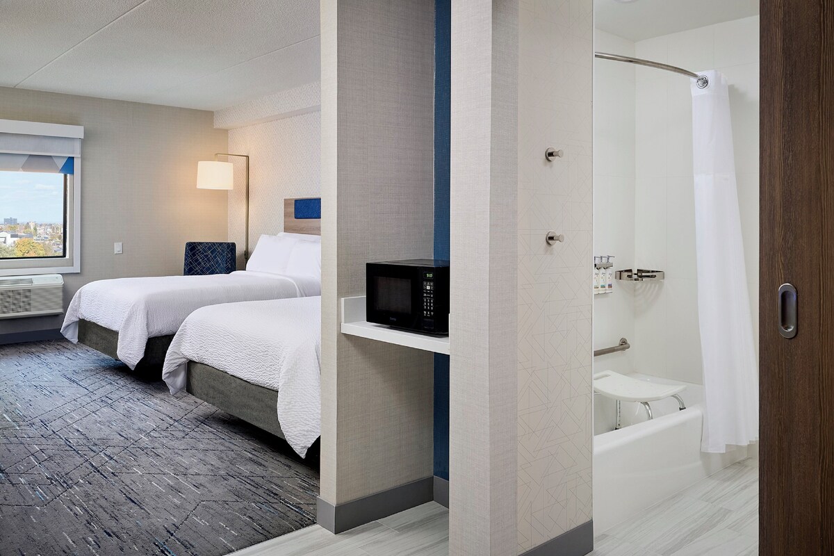 A hotel room with two beds, a microwave, a shower, and a window with a city view.