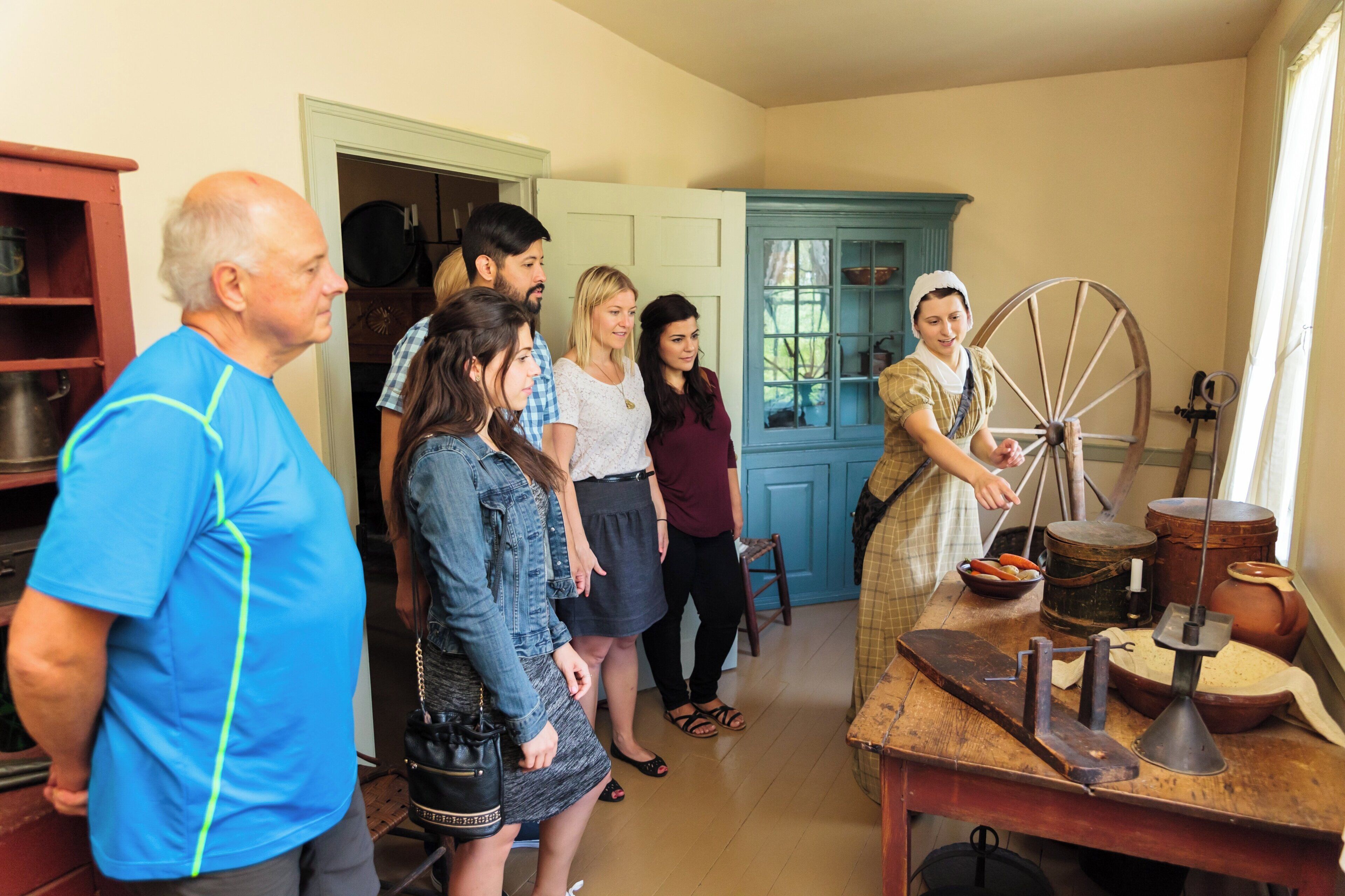 A group of people in a room with a spinning wheel, a wooden table with various items, and a blue cabinet.