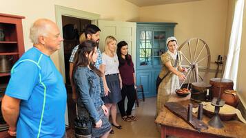 A group of people in a room with a spinning wheel, a wooden table with various items, and a blue cabinet.