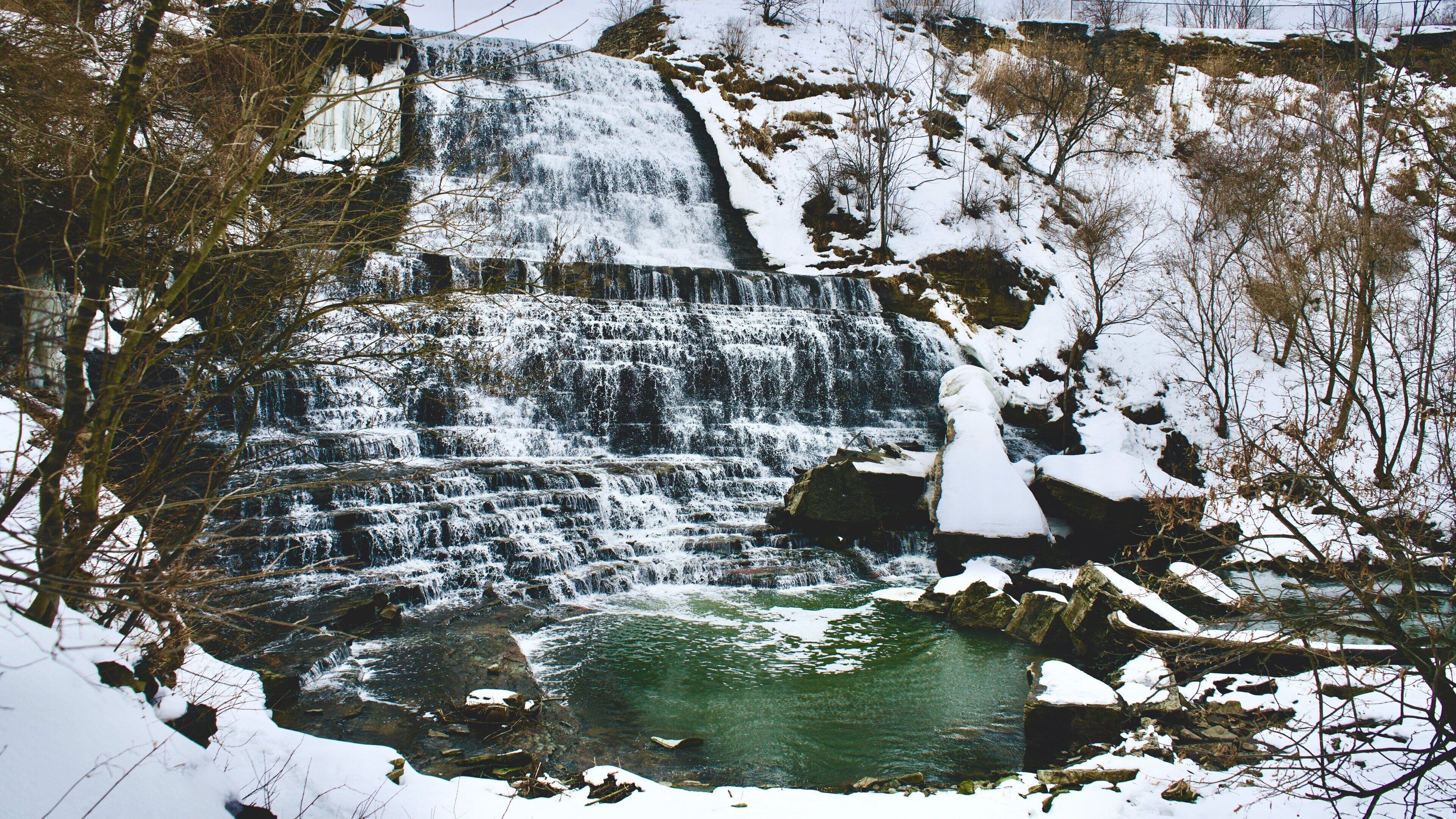 A waterfall cascading over rocks into a clear pool surrounded by snow-covered trees.