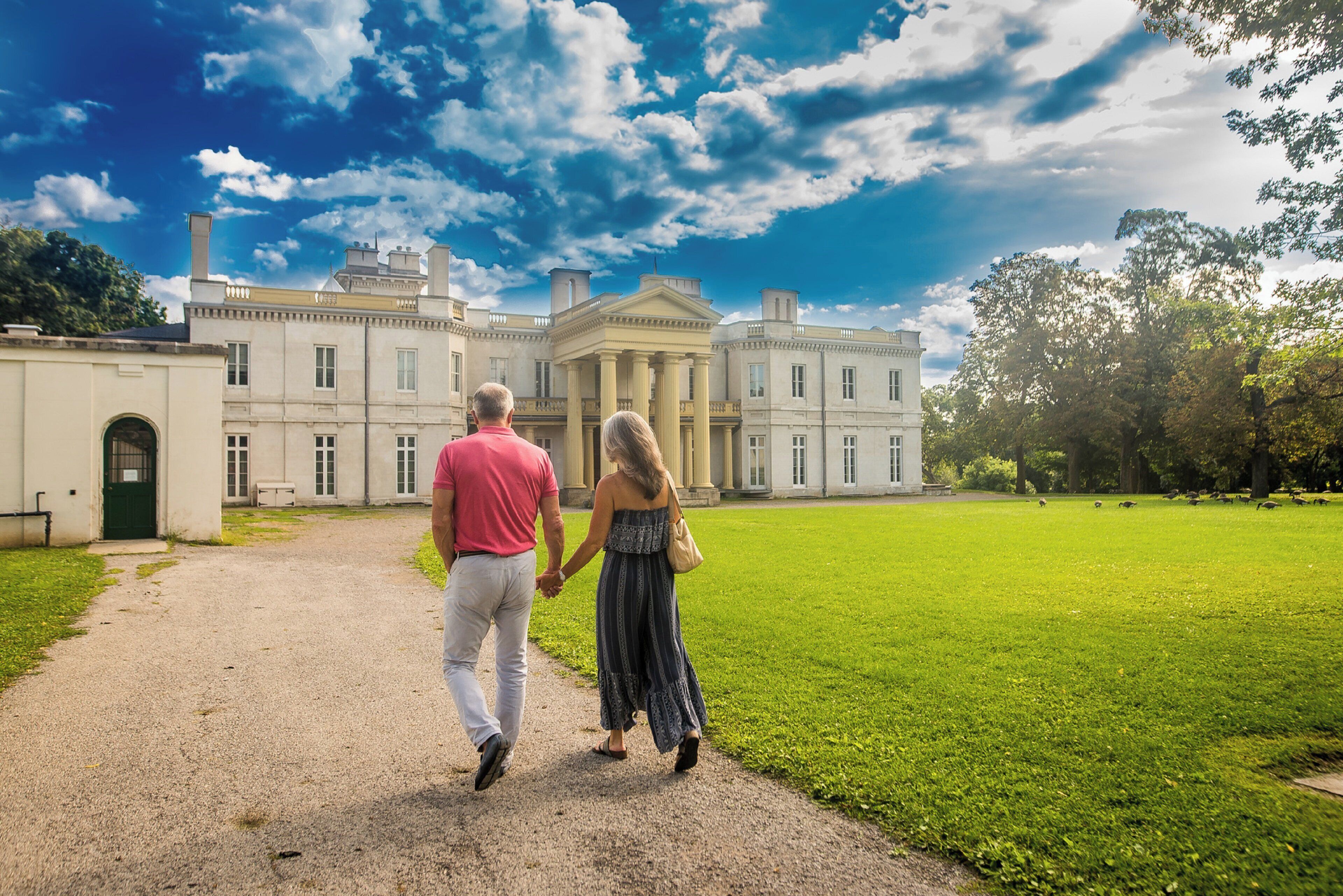 A couple walking on a gravel path towards a large, classical building with columns, surrounded by a well-maintained lawn.