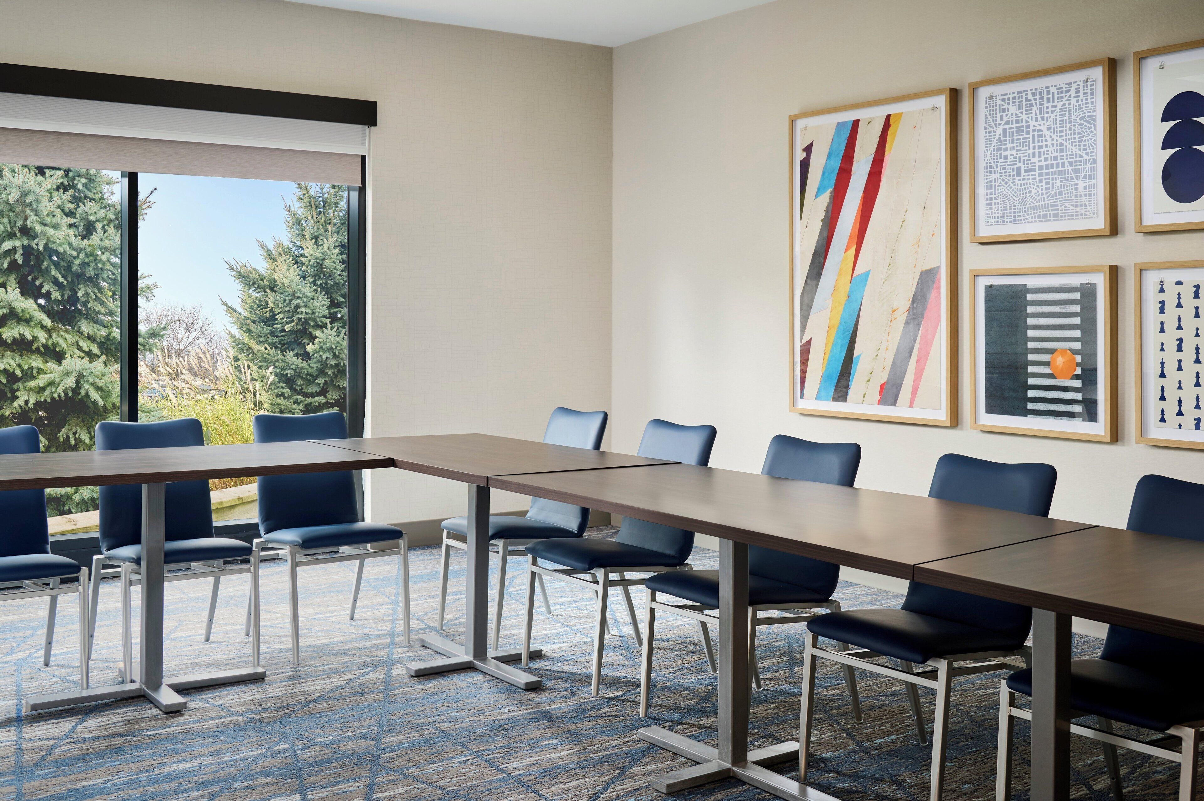 A conference room with a long table and blue chairs, framed artwork on the wall, and a view of trees through a large window.