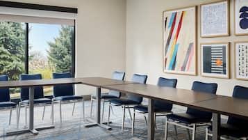 A conference room with a long table and blue chairs, framed artwork on the wall, and a view of trees through a large window.