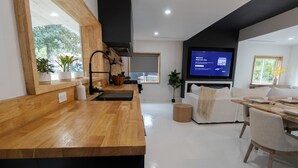 Una sala moderna con una mesa de comedor de madera, un sofá, un televisor y una zona de cocina con mostrador de madera y plantas en macetas.